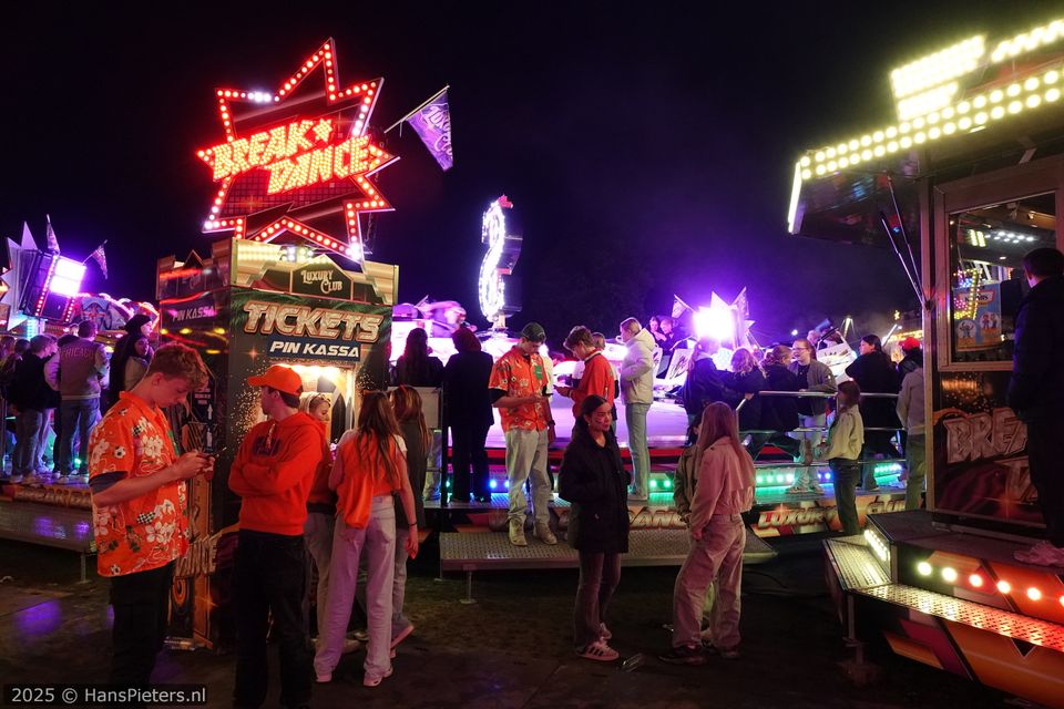 Kermisattractie Break Dance tijdens Koningsnacht in Woerden, Groene Hart, bezoekers bij de ticketkassa en jongeren op het verlichte draaiende kermispodium met neonreclame en avondverlichting op het feestterrein.