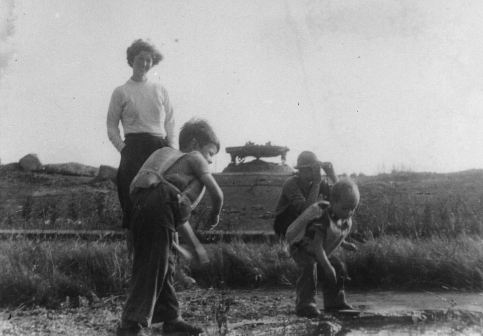 Een zeer oude foto van twee kinderen in zwemvesten bij het draaiplateau. Op de achtergrond kijkt de moeder toe, de vader zit gehurkt terwijl hij met zijn verrekijker naar het vasteland tuurt.