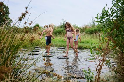 Een jongen en twee meisjes die in hun zwemkleding over ronde vlonders in het water lopen op het blotevoetenpad de Hege Gerzen.