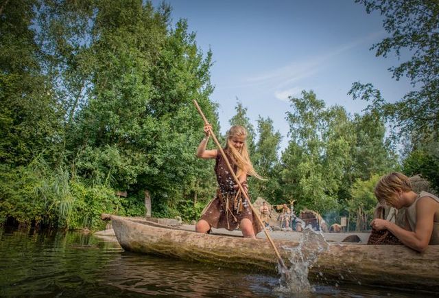 Kinderen in prehistorische kleding varen in een boomstamkano over het water in Archeon, omringd door groen en historische hutten in het Groene Hart.