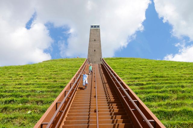 Kinderen lopen over de lange trap naar de uitkijktoren boven op de Pyramide van Austerlitz in Woudenberg. De groene piramide steekt af tegen een blauwe lucht met witte wolken.