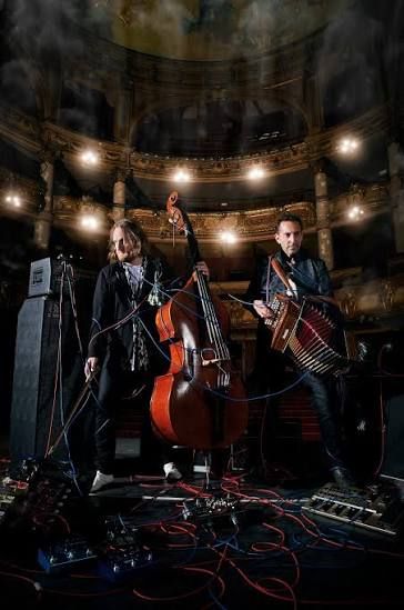 Donkere foto van twee mannen in het theater, de linker man houdt een contrabas vast en de rechter man een trekharmonica. Ze zijn omwikkeld met blauwe electriciteitsdraden.