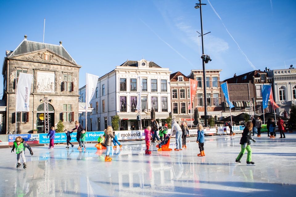 Een levendig winters tafereel op een zonnig plein: kinderen glijden en oefenen hun eerste rondjes op de ijsbaan, terwijl ouders en begeleiders toekijken. De historische gevels rondom het plein vormen een sfeervol decor en geven het schaatsplezier een typisch Hollandse uitstraling, waar stadse gezelligheid en winterse vrolijkheid samenkomen.