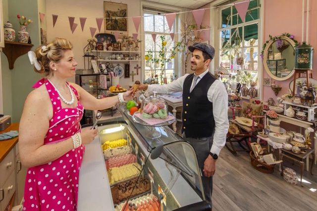 Een vrouw in retro kleding overhandigt een ijshoorntje aan een man in een winkel vol vintage spullen.
