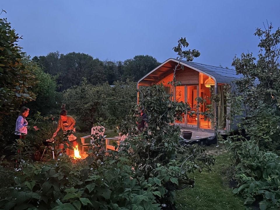 Avondbeeld van een sfeervol wikkelhuisje bij Proeftuin van Linschoten, met warm verlichte veranda en mensen rond een knisperend kampvuur tussen de bomen en struiken.