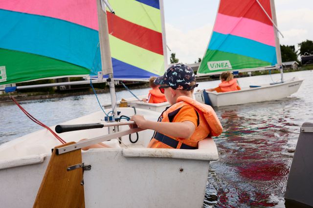 Drie kinderen die achter elkaar aan zeilen in een optimist met vel gekleurde zeilen op het Nannewiid.