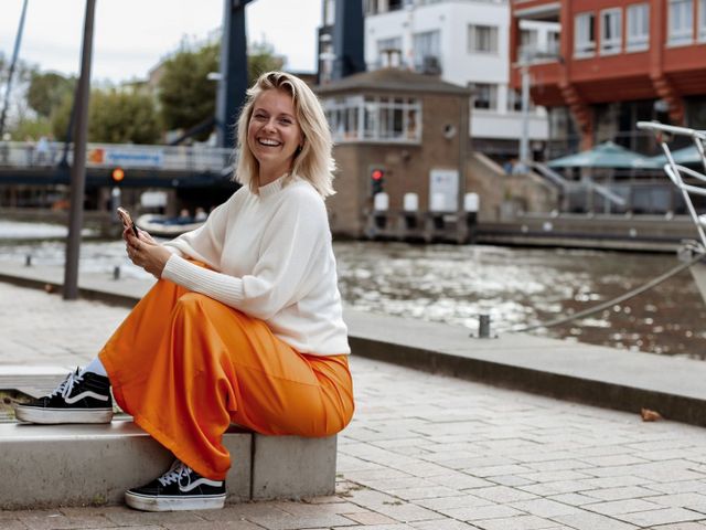 Vera de content & social media marketeer die op een straat verhoging zit met haar telefoon in d'r hand met op de achtergrond de Alphense brug.