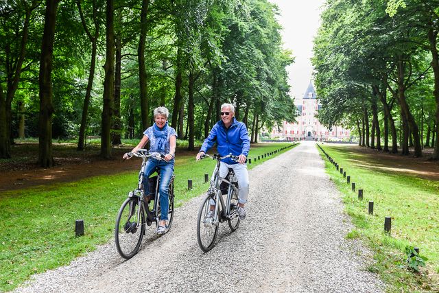 Twee oudere volwassenen fietsen lachend over een boomrijke oprijlaan van Kasteel de Haar in Haarzuilens.