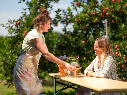 Een vrouw zit aan een tafel in een boomgaard, een andere vrouw serveert haar een kopje koffie.