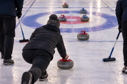 Mensen in winterkleding spelen recreatief curling op een tijdelijke ijsbaan. Vanaf de achterkant is te zien hoe een speler een curlingsteen richting het rood-blauwe doel schuift, terwijl andere spelers met bezems klaarstaan.