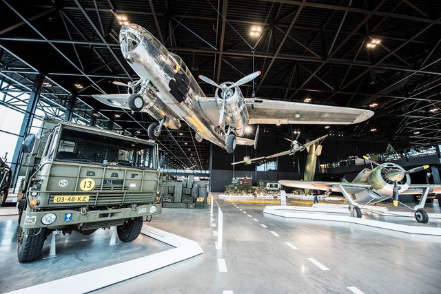 Overzicht van de ruime museumhal van het Nationaal Militair Museum met historische vliegtuigen en een militair voertuig. De tentoongestelde objecten geven een beeld van de Nederlandse militaire geschiedenis.