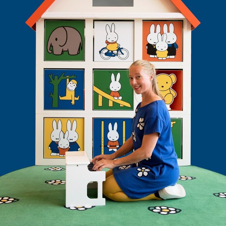 Foto van een blonde actrice in een blauwe jurk. Ze zit op haar knieën achter een klein wit pianootje voor een poppenhuis met allerlei verschillende afbeeldingen van Nijntje.