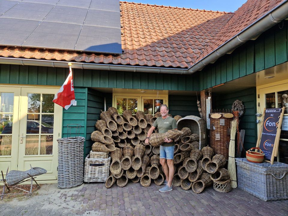 Ambachtelijke mandenmakerij De Broedkorf in Oudewater, Groene Hart, eigenaar staat buiten bij werkplaats met grote stapel handgemaakte rieten manden en vlechtwerk van wilgentakken.