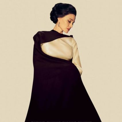 Foto van een vrouw in een crème-kleurige jurk met een zwarte cape. Ze heeft donker opgestoken haar en staat voor een crème-kleurige achtergrond.