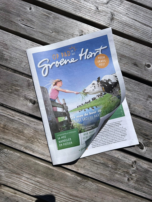 Magazine Op Pad in het Groene Hart, recreatiekrant van Stichting Groene Hart, Groene Hart. Tijdschrift met inspiratie voor wandelen, fietsen en uitjes ligt op een houten picknicktafel in de buitenlucht.