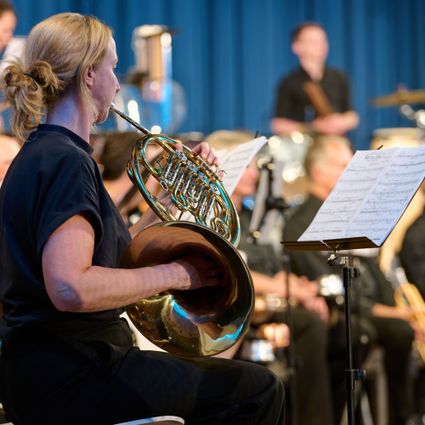 Foto van een dame die blaast op een hoorn achter een muziekstandaard met bladmuziek daarop.