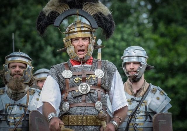 Romeinse centurio in harnas met helm en onderscheidingstekens geeft bevel tijdens een historische re-enactment in Archeon, met soldaten op de achtergrond.
