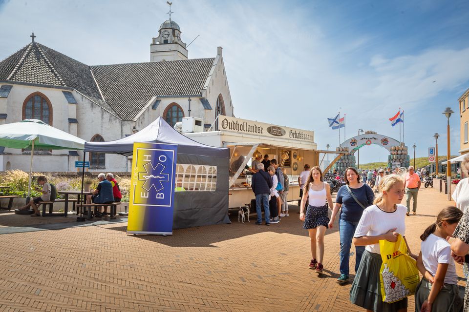 Bezoekers struinen over de toeristenmarkt in Katwijk langs een oliebollenkraam, met op de achtergrond de Andreaskerk, ook wel Witte kerk genaamd.