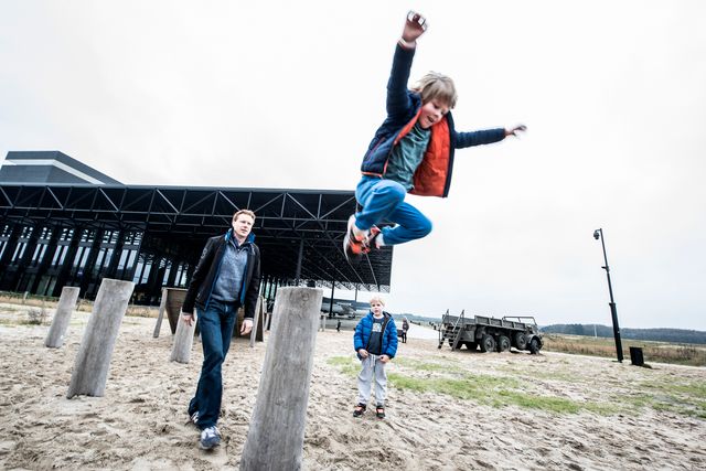 Een kind springt tussen de houten speeltoestellen bij het Nationaal Militair Museum in Soest. Op de achtergrond zijn andere bezoekers, een militair voertuig en het museumgebouw te zien.