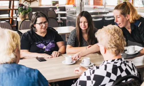 Vier vrouwen zitten aan een tafel, een vijfde vrouw serveert koffie.