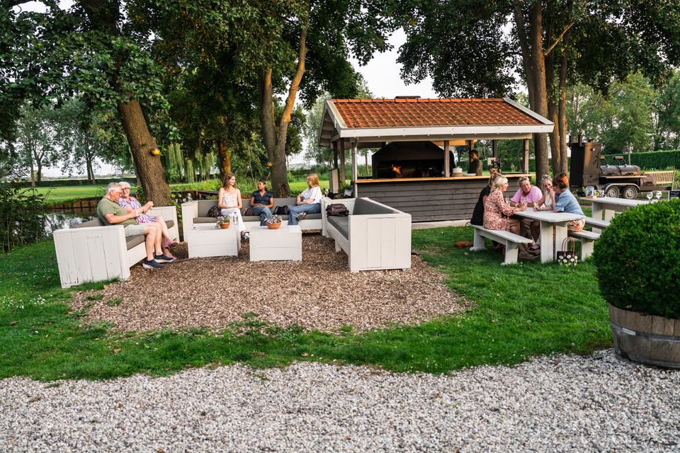 Gezellige groepen mensen op het braai-terras van Buitenplaats Kameryck, zittend aan picknicktafels en loungebanken in het groene landschap.