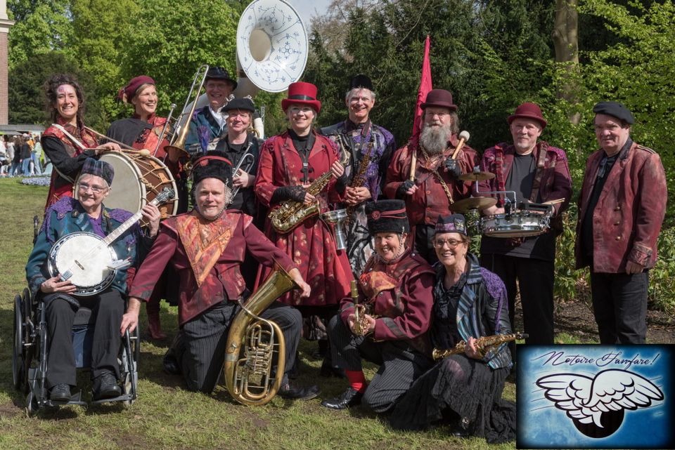 Foto van dames en heren van de fanfare met paarse en donkerrode kleding en diverse muziekinstrumenten. Ze staan in de openlucht in een bosrijke omgeving.