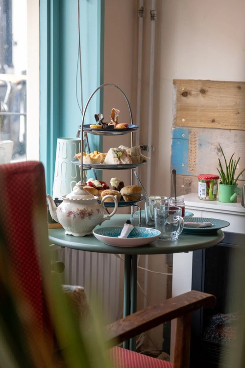 Een tafel gedekt met een etagere vol lekkere hapjes, een theepot en glazen.