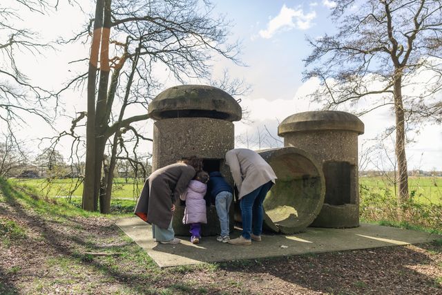Een gezin met lange jassen aan kijkt in een buis in een groen omgeven Grebbelinie.
