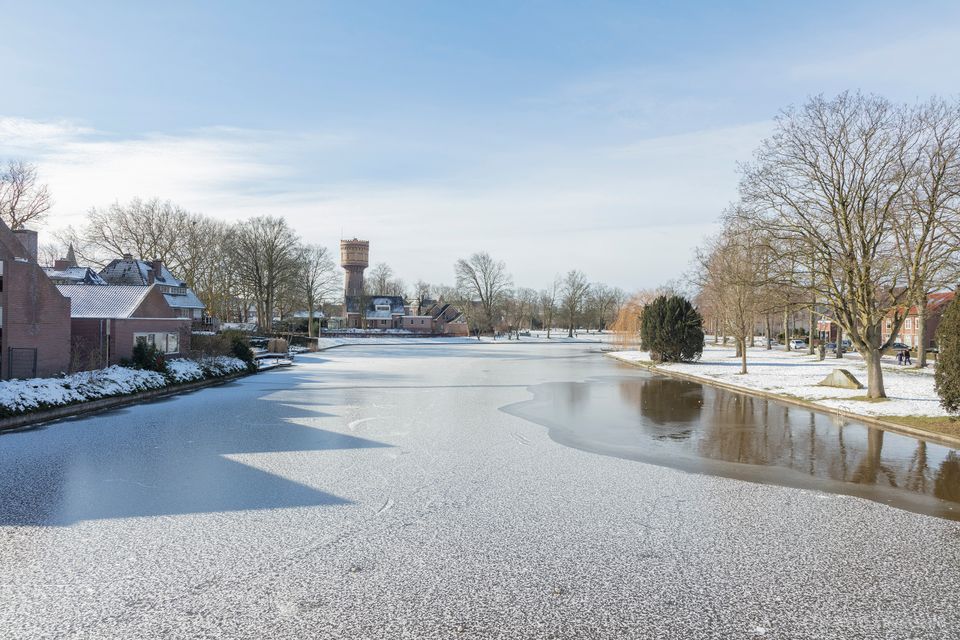 Een serene winterdag in Woerden, waar een dun laagje ijs het water bedekt en de iconische watertoren statig boven de met sneeuw bestoven huizen en bomen uittorent. Het zachte winterlicht en de rustige sfeer geven het landschap een bijna schilderachtige kwaliteit.