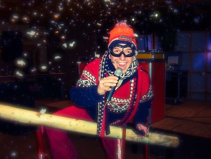 Persoon in vrolijke winteroutfit met rode muts, sjaal en trui zingt enthousiast in een microfoon terwijl sneeuwvlokken door de lucht dwarrelen. Hij leunt over een houten hek op een sfeervol verlicht podium tijdens een après-ski party in winterse sfeer.