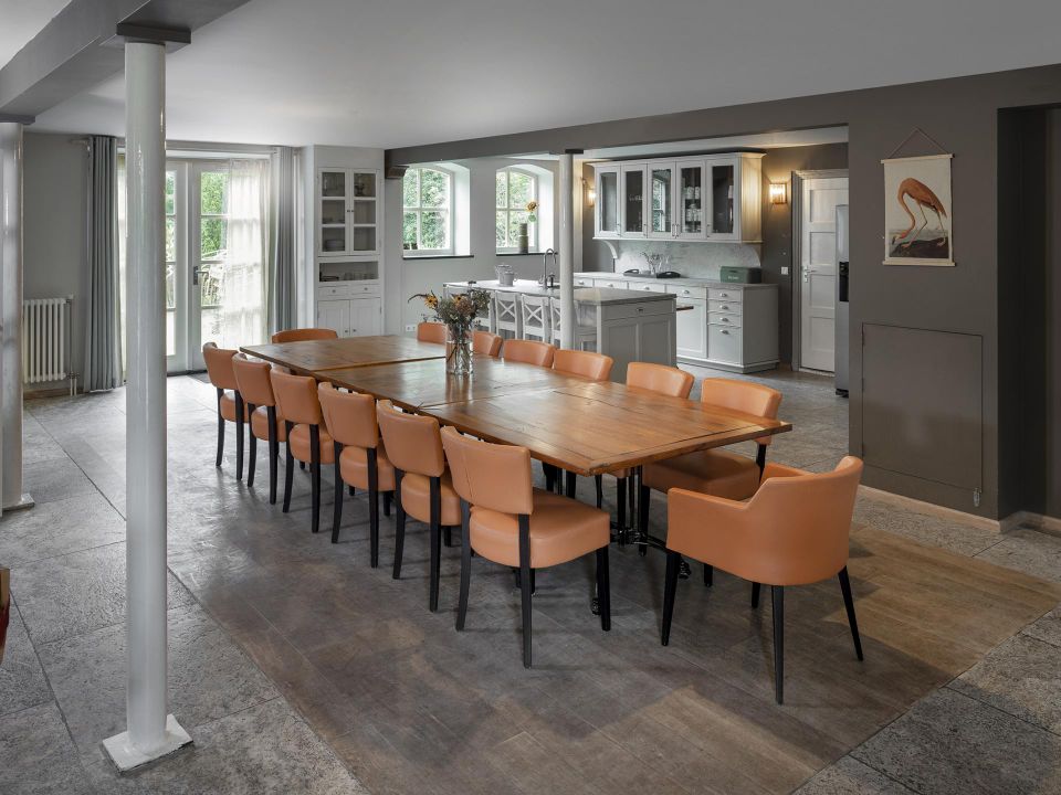 Zaal met lange eettafel en 14 oranje kuipstoelen en op de achtergrond een witte buffetkeukenblok.