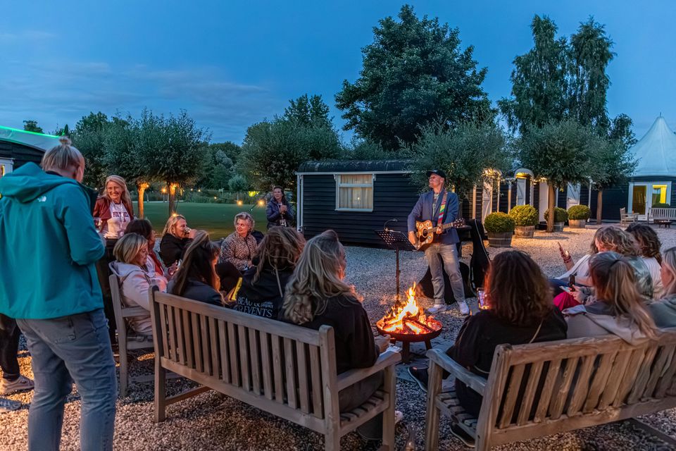 Groep mensen zit rond een kampvuur bij de Veldhuysjes van Kameryck terwijl een muzikant live gitaar speelt op een sfeervolle zomeravond.