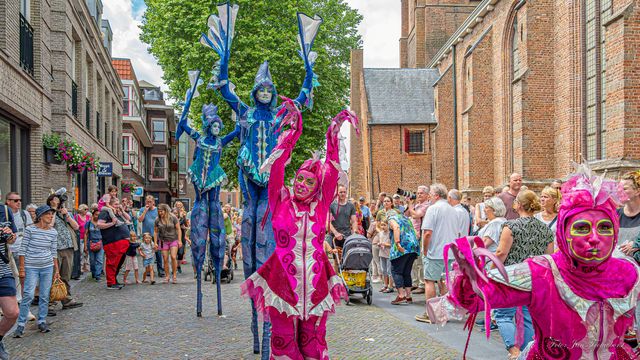 Kleurrijk straattheater in het centrum van Woerden, regio Groene Hart, met steltenlopers en theaterartiesten tijdens een zomers evenement tussen publiek en historische gebouwen.