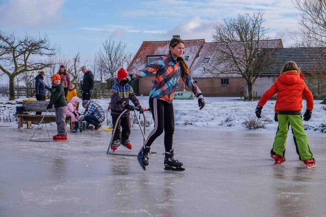 Een vrolijk wintertafereel op het platteland, waar kinderen en families genieten van een middag schaatsen op natuurijs. Met boerderijen op de achtergrond, kleurige winterjassen en enthousiaste beginners op steunrekjes ademt dit beeld pure Hollandse gezelligheid — een echte winterdag zoals die bedoeld is.