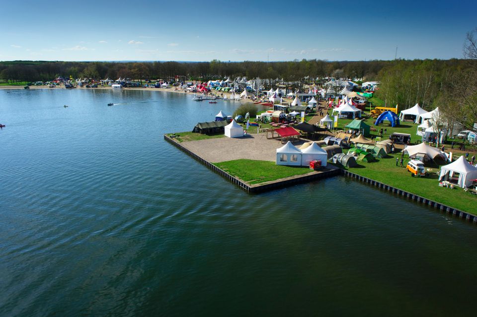 Een luchtfoto van een festivalterrein aan het water, met veel witte tenten en kramen.