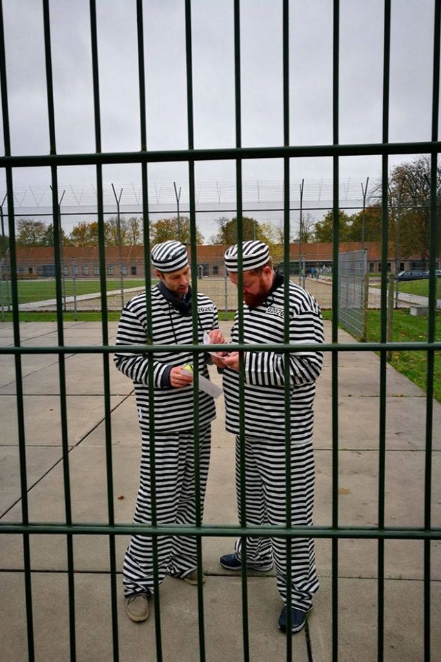 Twee mannen in een boevenpak staan achter een hek naar een papiertje te kijken.
