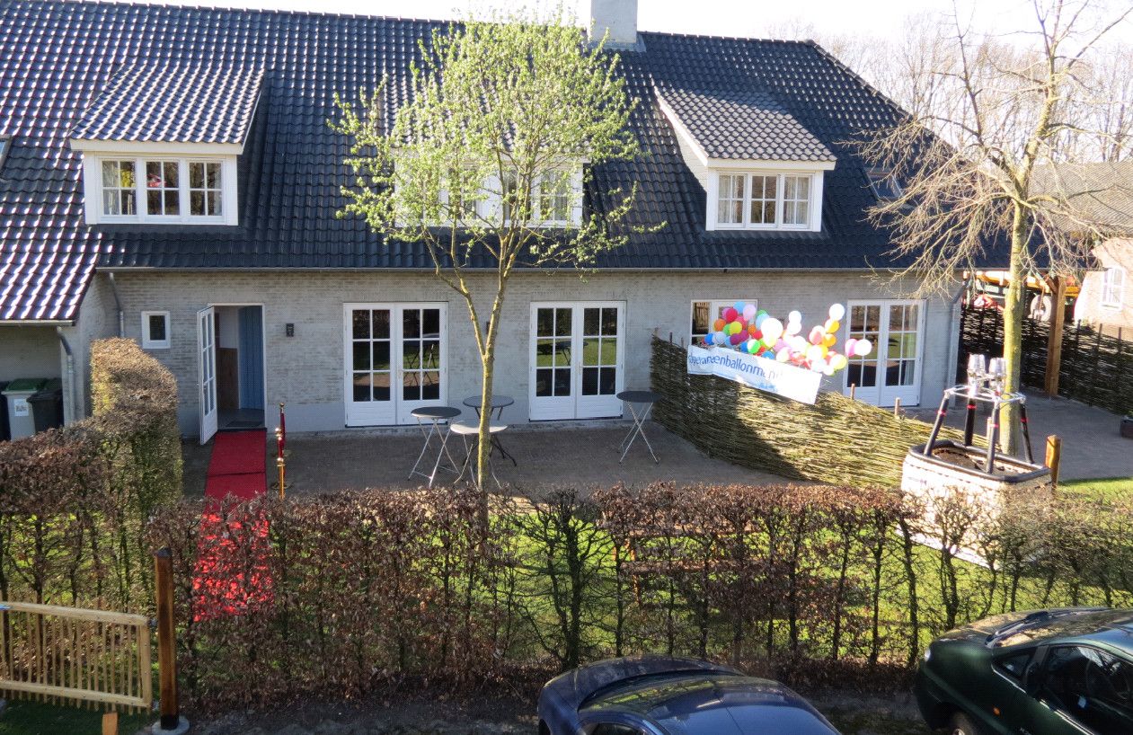 Foto van lichtgrijze boerderij met zwart pannendak en dakkapellen en een tuin met beukenhaag ervoor.