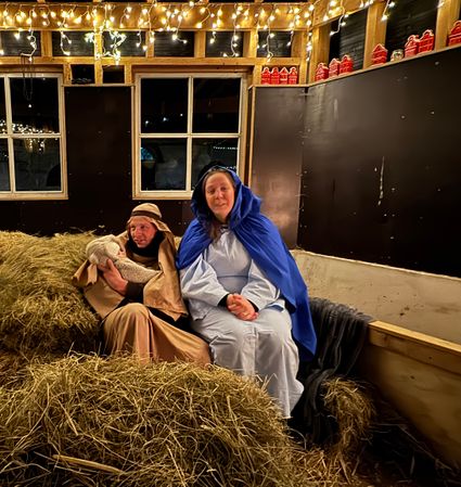In een warme kerststal zitten twee deelnemers verkleed als Jozef en Maria tussen het stro, met zachte kerstverlichting boven hen. Jozef houdt een babypop vast terwijl Maria naast hem zit in een sfeervolle setting.