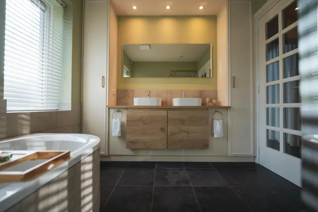 Badkamer met ligbad, dubbele wastafelmeubel met grote spiegel, donkere vloertegels en glazen schuifdeur.