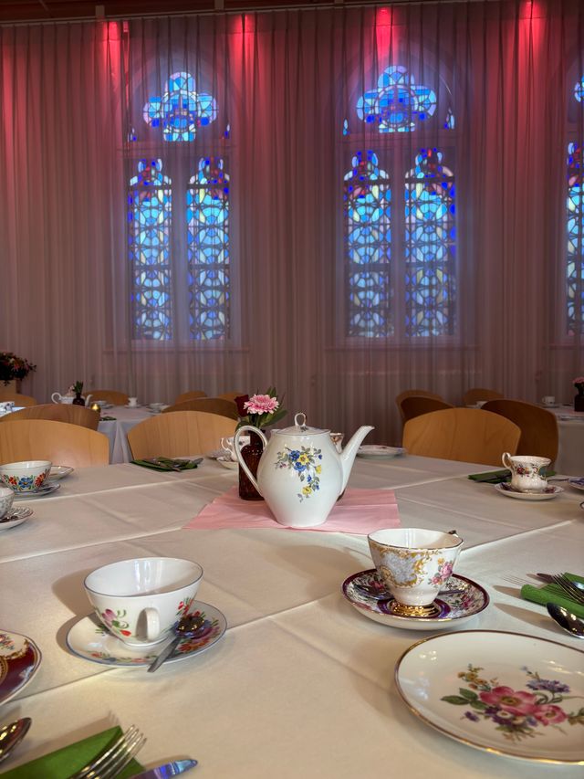 Gedekte tafel met theekopjes, schotels, lepels en servetten; in het midden staat een theepot en klein bloemvaasje. Andere tafels zijn zichtbaar in de zaal.