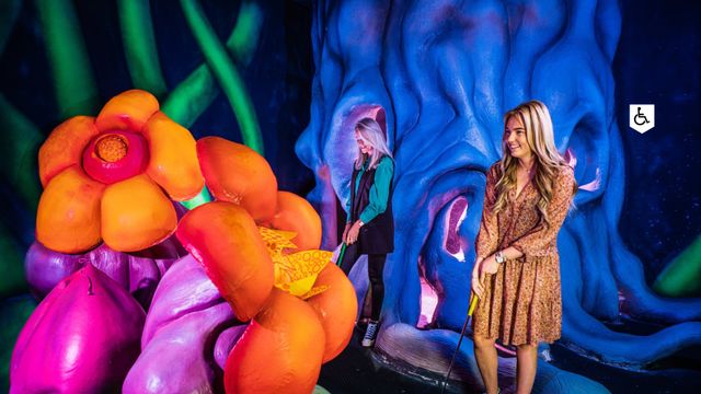 Uitgaanslocatie De Fabriek Woerden, provincie Utrecht, Groene Hart, twee bezoekers spelen glow in the dark minigolf tussen felgekleurde bloemen en fantasierijke decorstukken in een indoor activiteitencentrum.