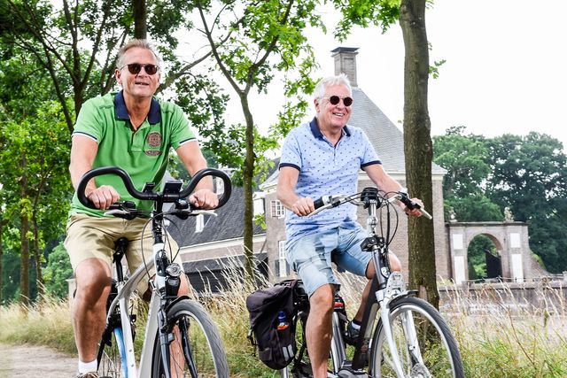 Twee mannen fietsen lachend door het groene landschap bij een historisch gebouw in de omgeving van Woerden.