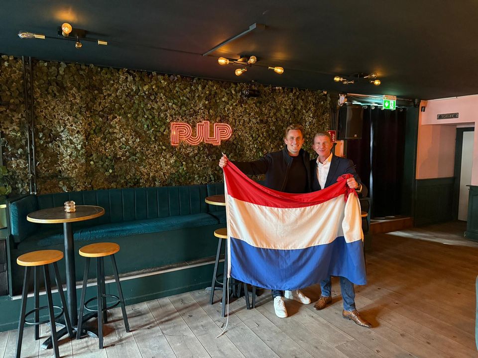 Burgemeester Jack de Vries (rechts) en Wiebe Kuiper, voorzitter van Koninklijk Horeca Nederland Maassluis (links), poseren samen met de Nederlandse vlag en onderstrepen de samenwerking rondom het WK Voetbal 2026.