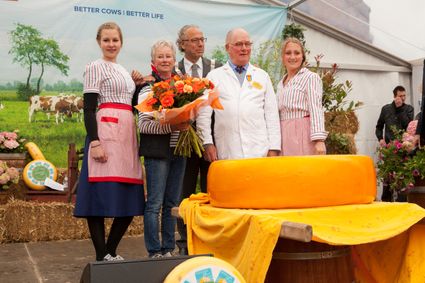 Kaasmarkt evenement Graskaasdag Woerden in het Groene Hart met prijsuitreiking rond een grote graskaas op houten vat, kaasmeester in witte jas, deelnemers in traditionele klederdracht en boeket bloemen voor decor van weiland met koeien.
