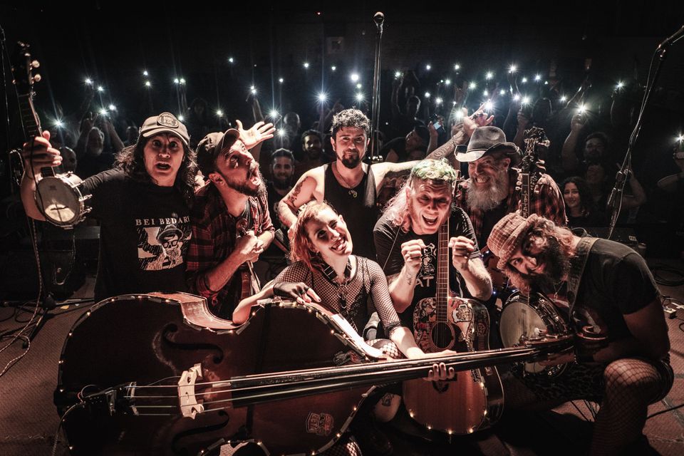 Groepsfoto van de band Angry Zeta op het podium, poserend met instrumenten voor een enthousiast publiek met oplichtende telefoons in de lucht.
