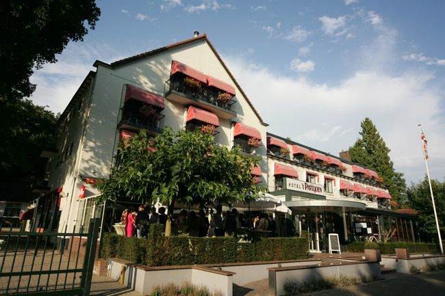 Buitenzijde van Hotel ’t Paviljoen in Rhenen, met rode zonneschermen en een terras met bezoekers. Het hotel ligt in een groene omgeving op de Utrechtse Heuvelrug.