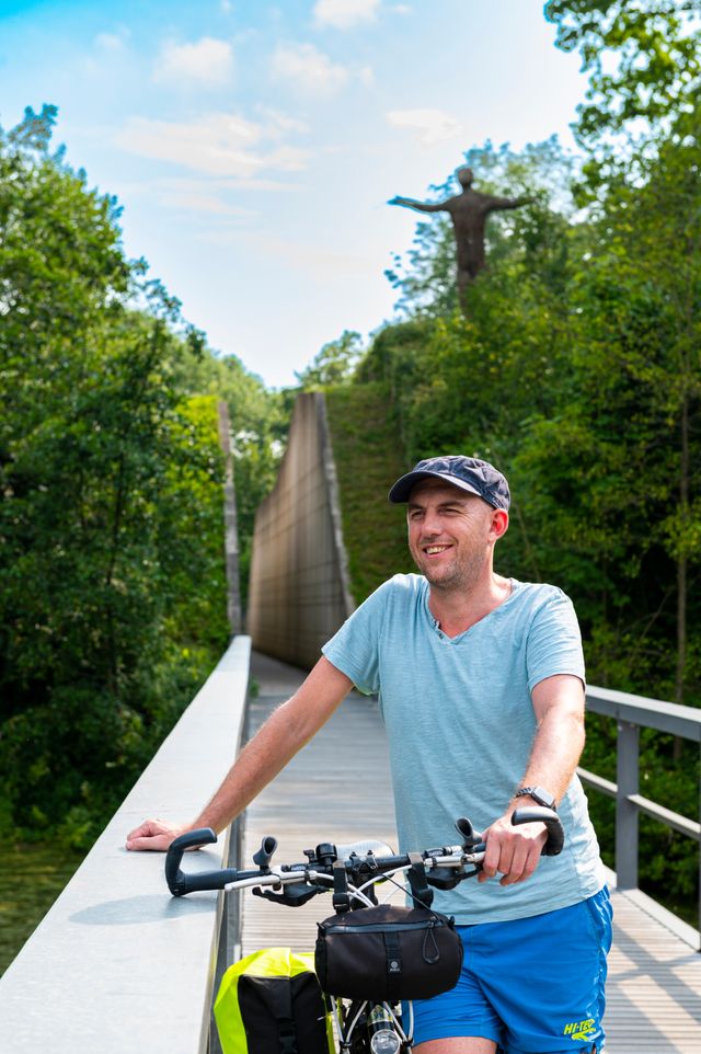 Een man met een fiets staat op een loopbrug die richting een aarden wal gaat, waarin een coupure is aangebracht. Bovenop de wal staat een kunstwerk van hout dat een mensfiguur voorstelt.
