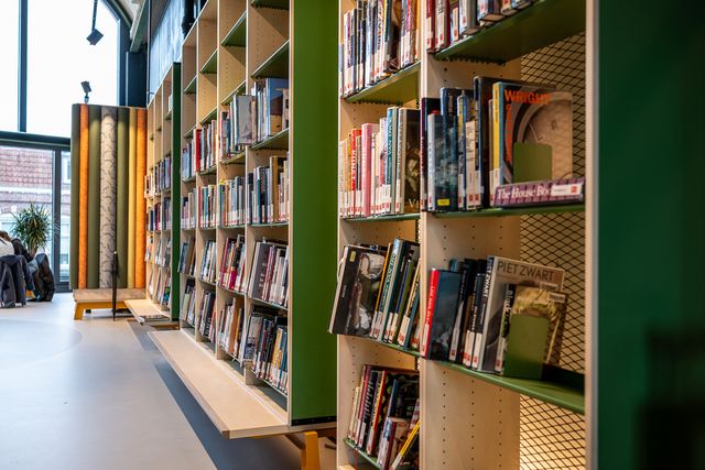 Lange rij boekenkasten met uitgestalde boeken in een bibliotheek, naast een looproute met zitplekken bij het raam, bedoeld voor het zoeken en lezen van boeken.