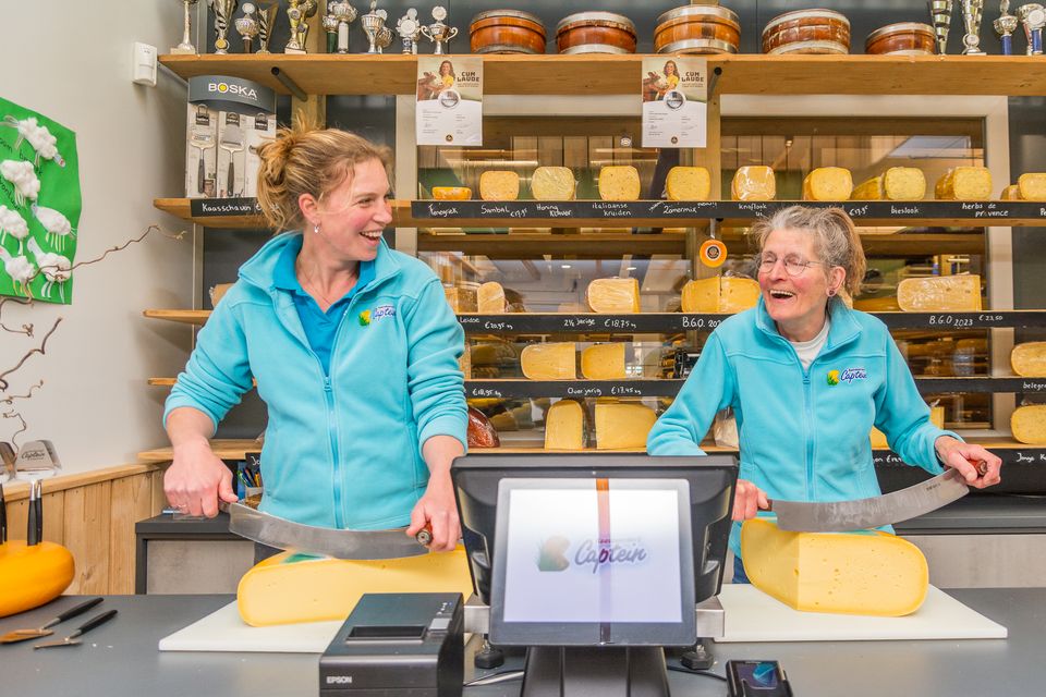 Twee vrouwelijke medewerkers staan lachend achter de toonbank kaas te snijden, in de kaaswinkel van Kaasboerderij Captein.