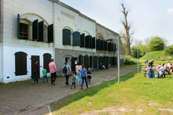 Een bakstenen fort. Voor het fort langs lopen twee gezinnen van in totaal acht personen. In het gras voor het fort zitten bezoekers op picknickbanken.